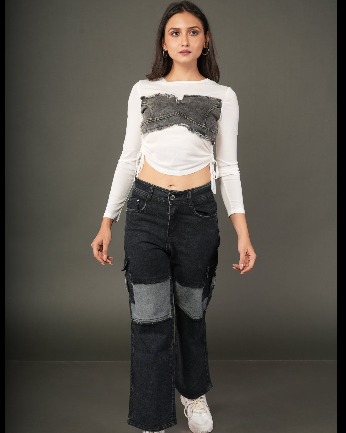 A woman modeling a white long-sleeve crop top with a gray patchwork chest panel and dark blue patchwork flare jeans, paired with white sneakers.