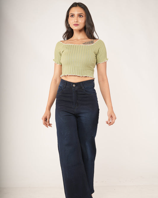 A woman modeling a light olive green ribbed short-sleeve crop top with a ruffled neckline, paired with dark blue jeans.