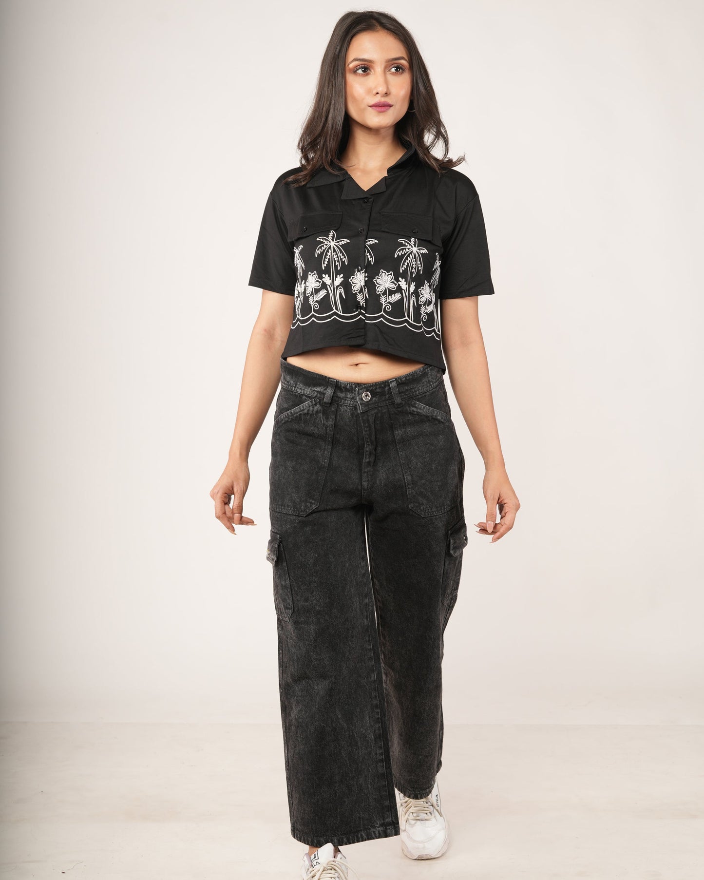 A woman modeling a black cropped t-shirt with a white palm tree graphic and Charcoal 6-Pocket Cargo wide-leg cargo jeans, standing against a plain light beige studio background.