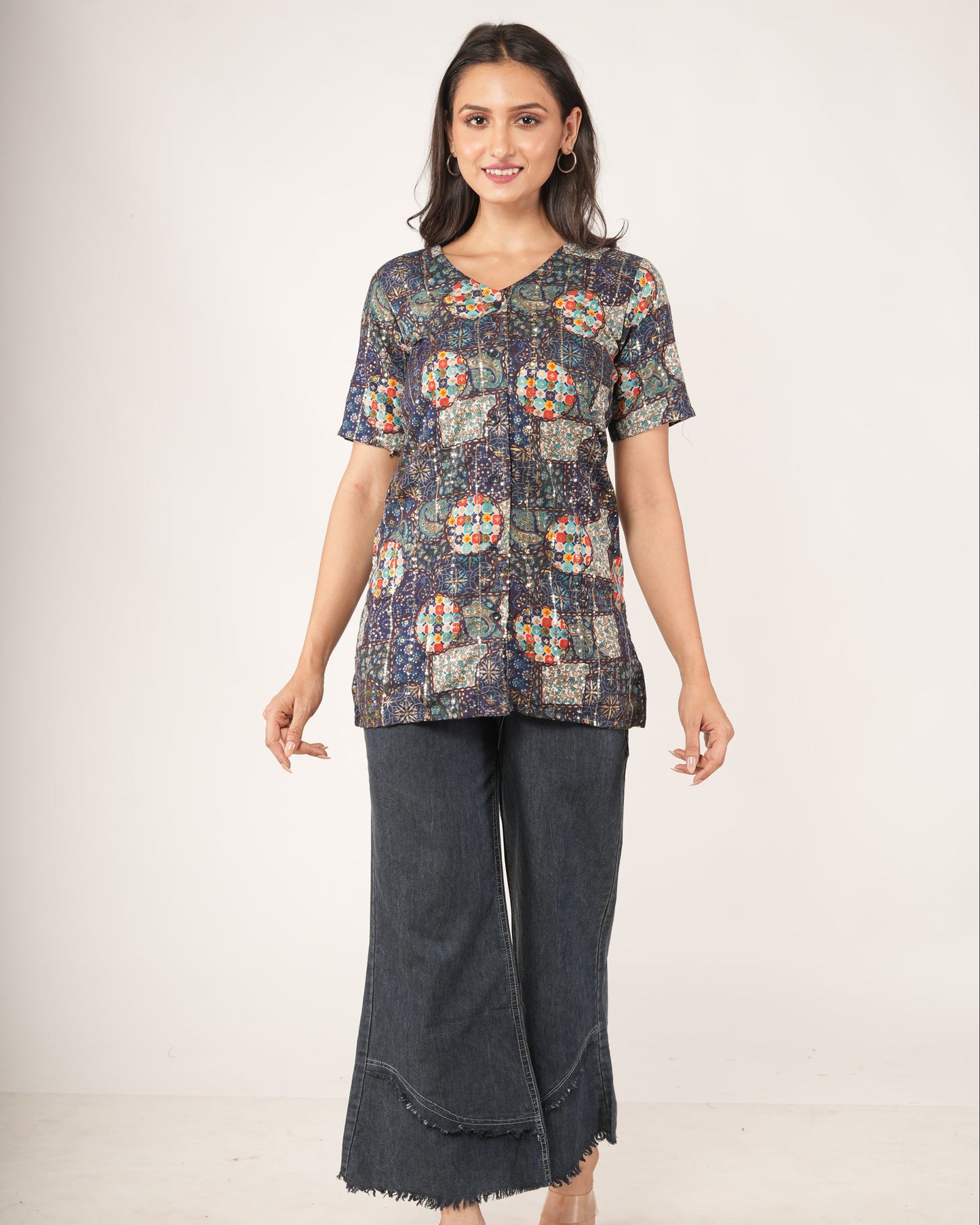 A woman modeling Ethnic Fusion Embroidered Sequin Top  with a multicolor paisley pattern on a dark base, paired with dark gray wide-leg jeans and clear high-heeled sandals, against a plain white studio background.