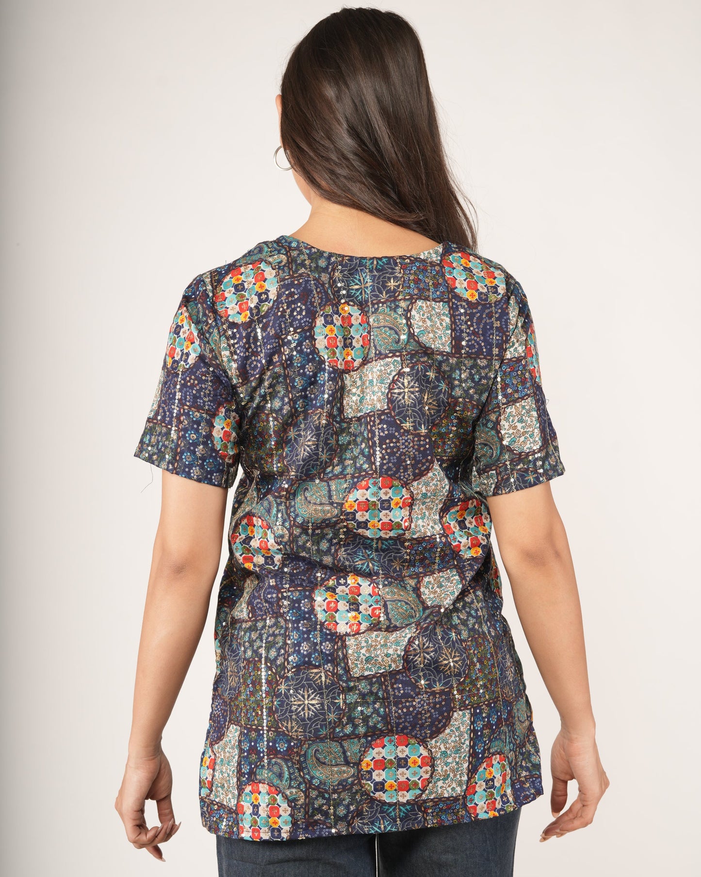A woman viewed from the back wearing a short-sleeve Ethnic Fusion Embroidered Sequin Top print in blue, beige, red, and green, paired with dark jeans.
