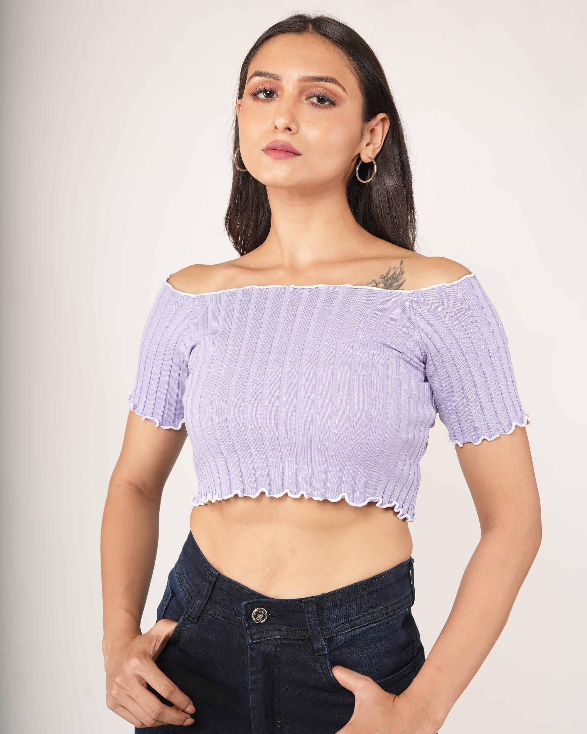 A woman modeling a lavender/light purple off-the-shoulder ribbed crop top with short sleeves and a scalloped hem, paired with dark blue jeans.