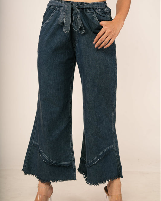 A model wearing dark blue wide-leg denim flare pants with a drawstring waist and frayed hems, paired with a patterned crop top and clear high-heeled sandals.
