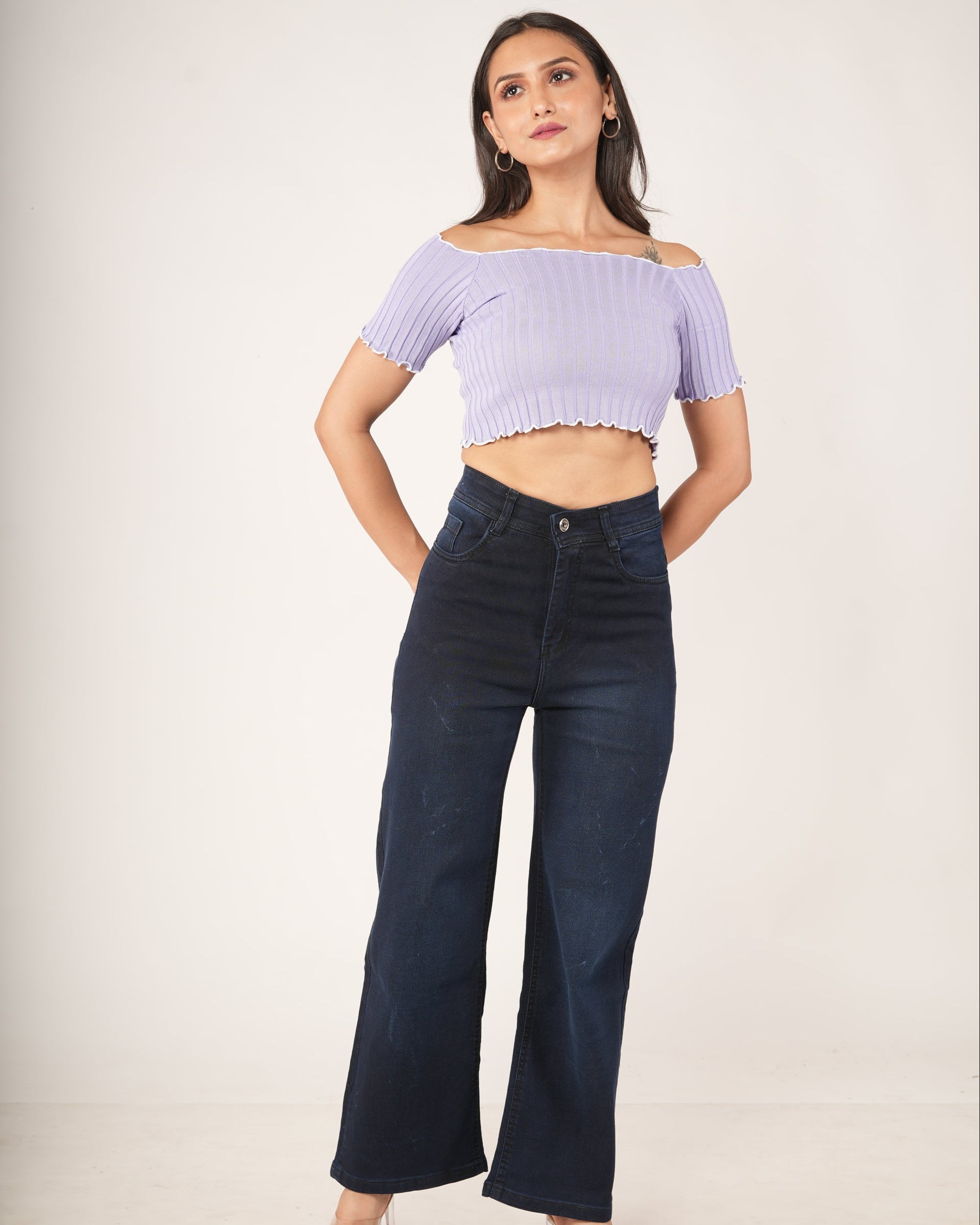 A woman modeling a light purple ribbed off-the-shoulder crop top and dark navy wide-leg jeans against a plain off-white studio background.
