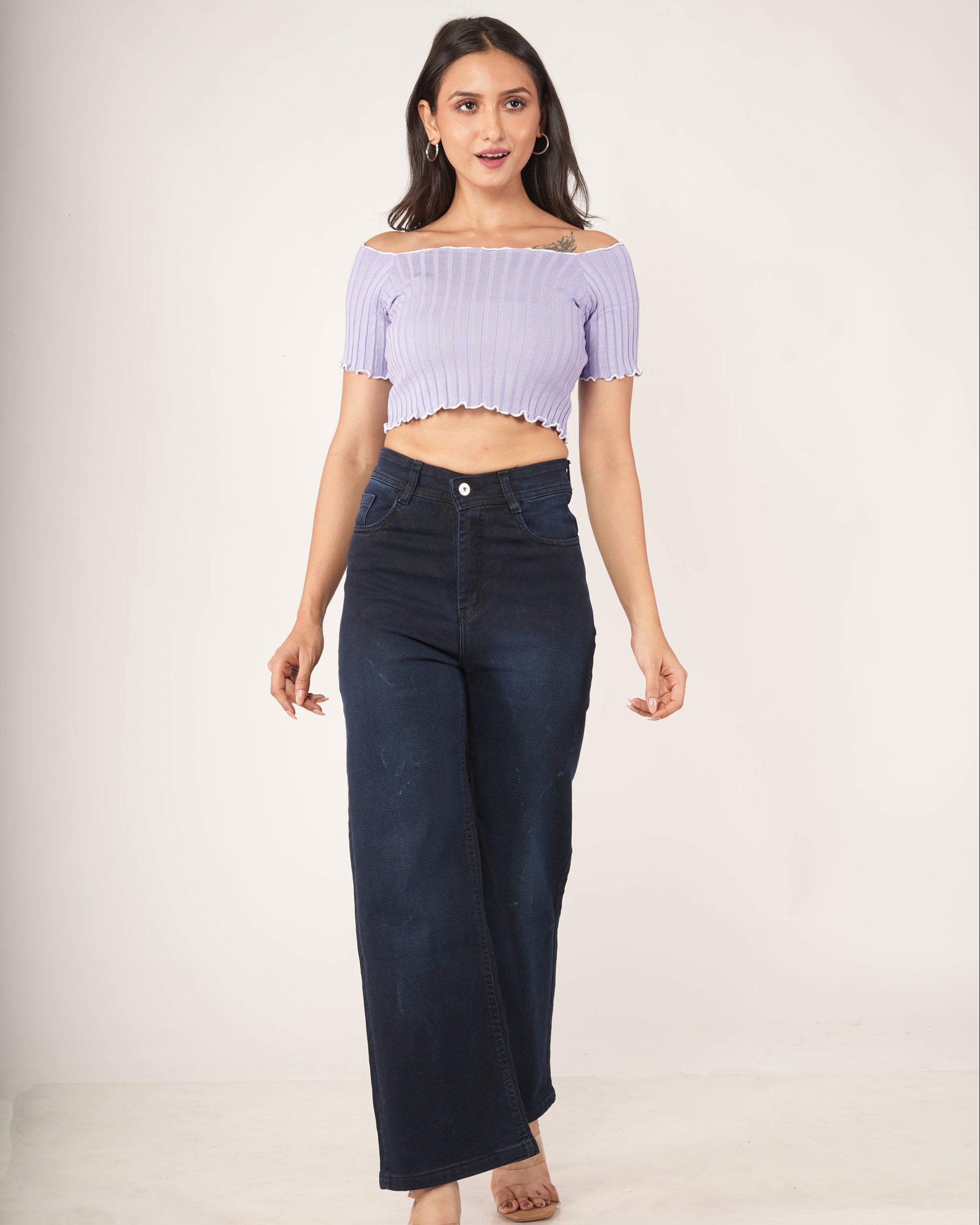 A woman modeling an off-the-shoulder light purple ribbed crop top and dark navy wide-leg jeans against a plain light gray studio background.