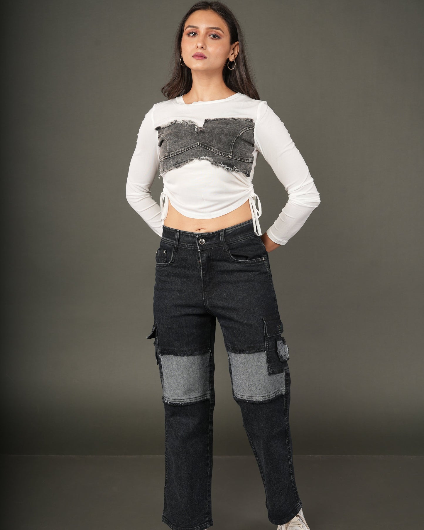 A model stands against a dark gray studio background wearing a white long-sleeve crop top with a gray textured chest panel and side drawstrings, paired with dark patchwork cargo jeans and white sneakers.