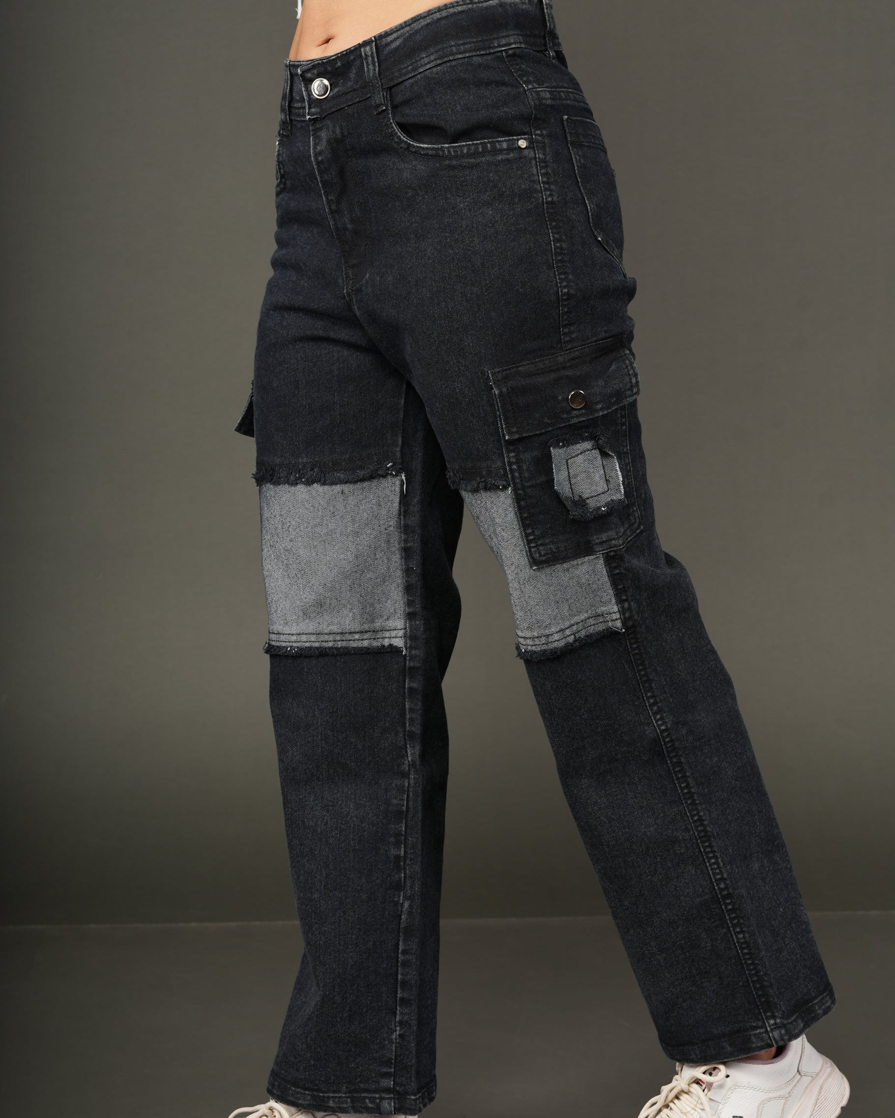 A person wearing dark patchwork cargo jeans with gray denim patches on the thighs and knees, styled with a white cropped top and white sneakers.