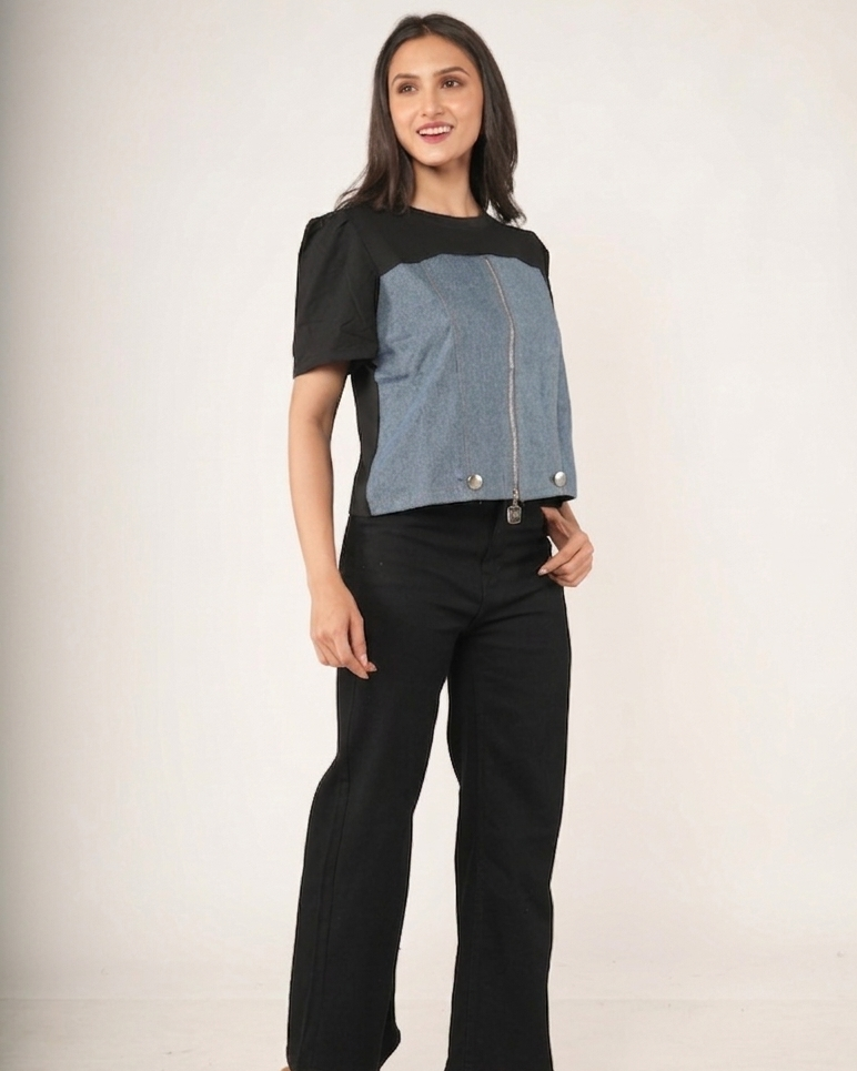 A woman modeling a color-blocked short-sleeve top with black sleeves and a blue denim-like front panel, paired with black wide-leg pants.