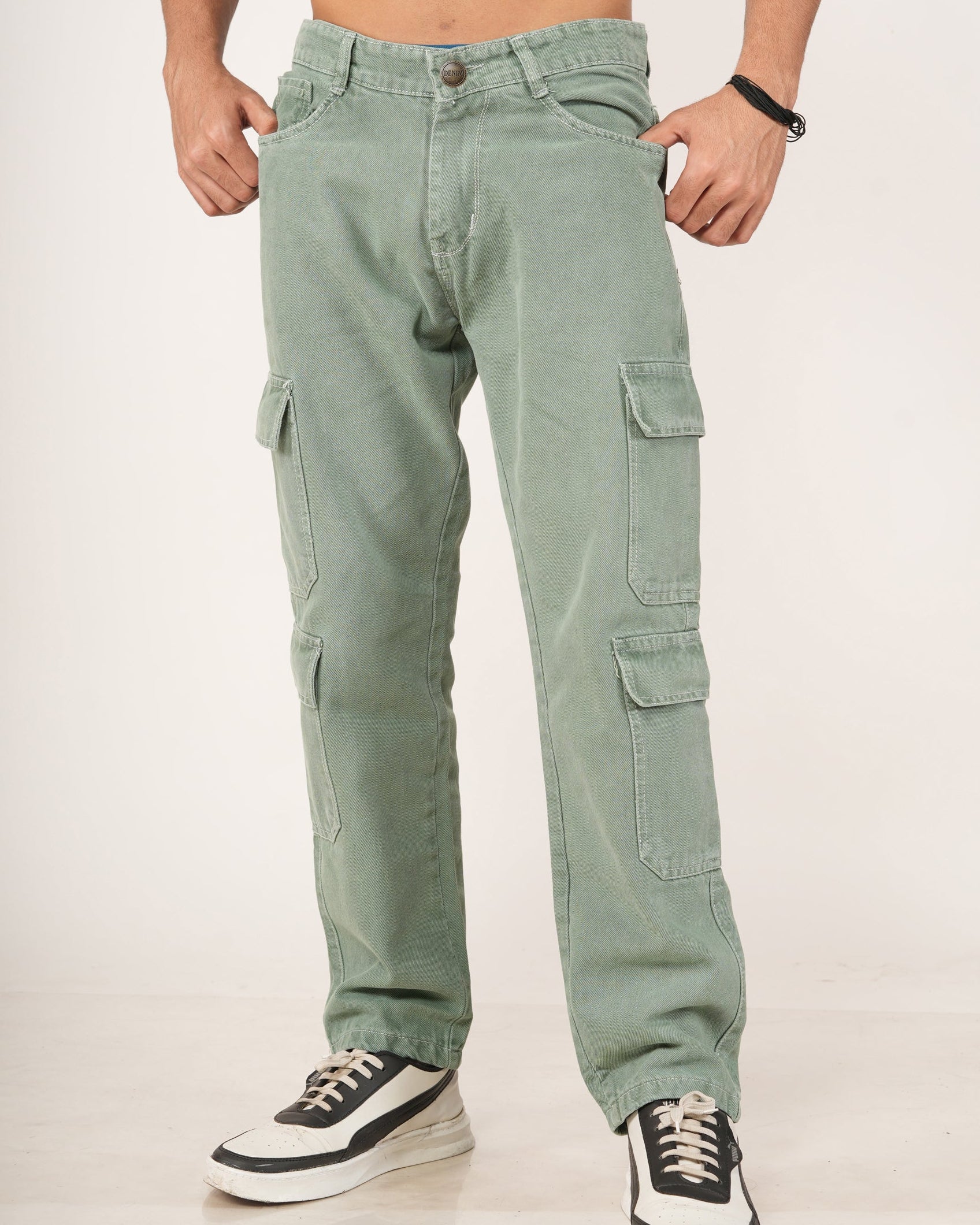 A man wearing sage green 8 pocket cargo jeans, shown from the chest down against a plain light background. He is wearing black and white sneakers.