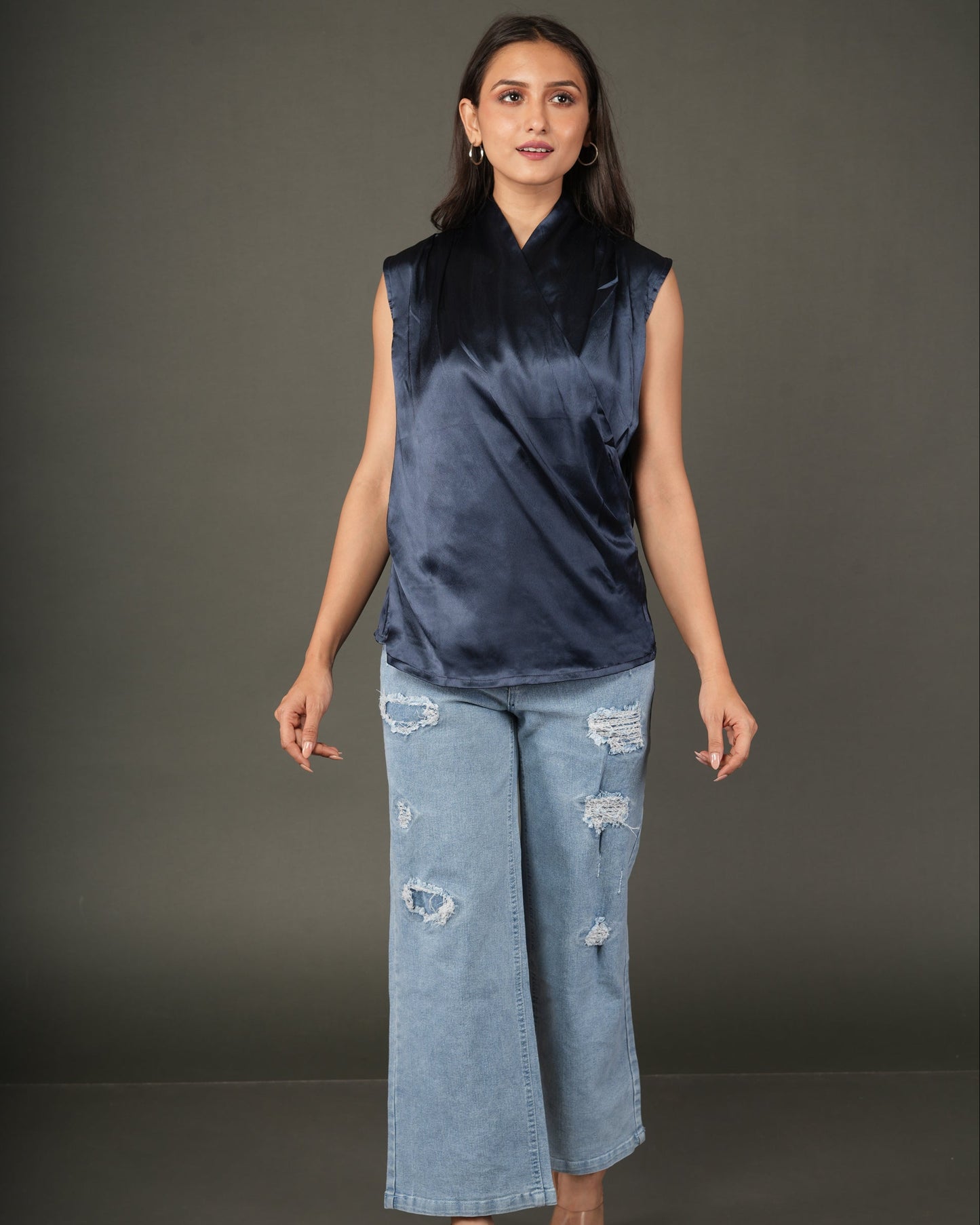 A woman modeling a sleeveless dark navy satin top with a high collar, paired with light-wash distressed wide-leg jeans and brown sandals.