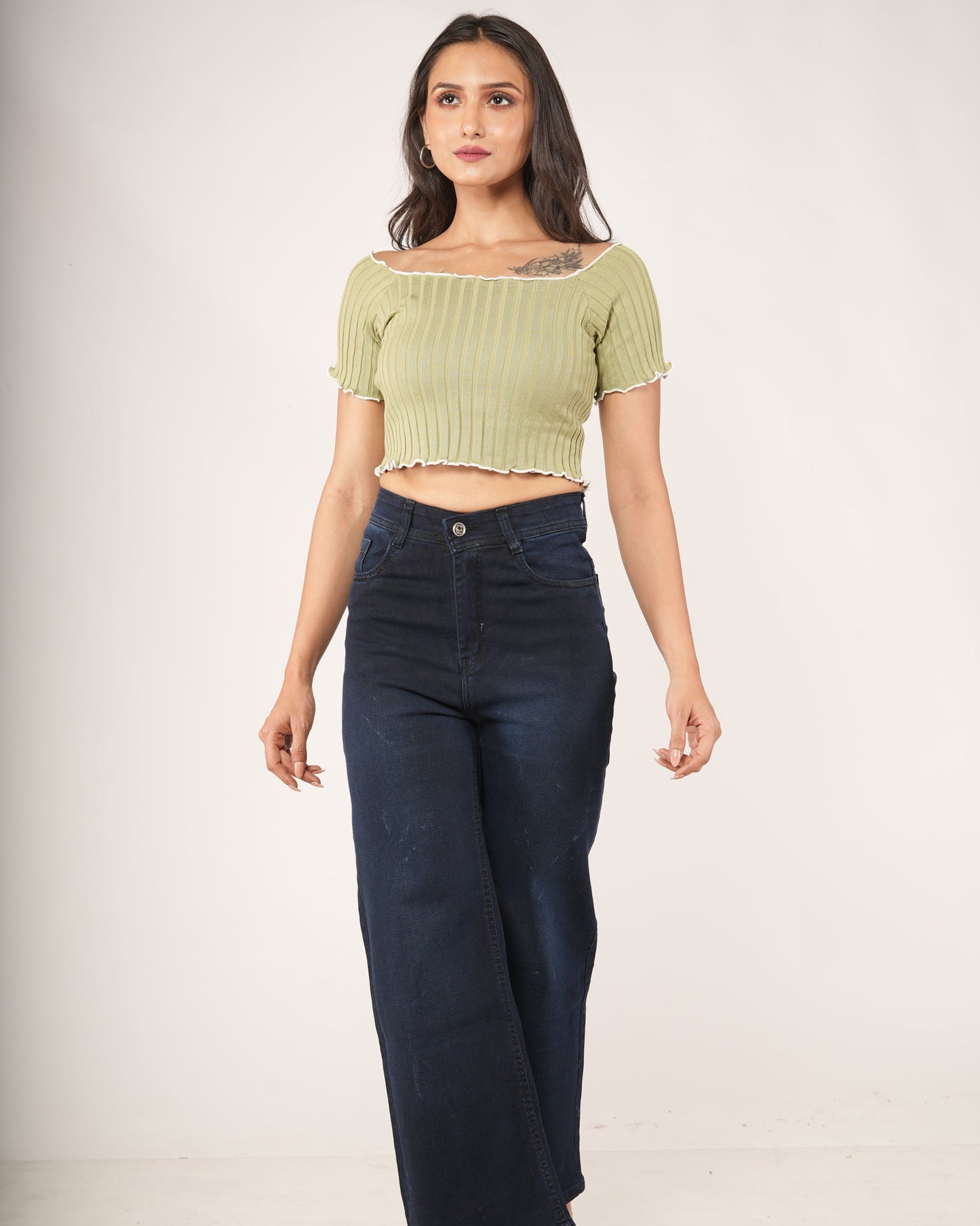 A woman modeling a light olive green ribbed short-sleeve crop top with a ruffled neckline, paired with dark blue jeans.