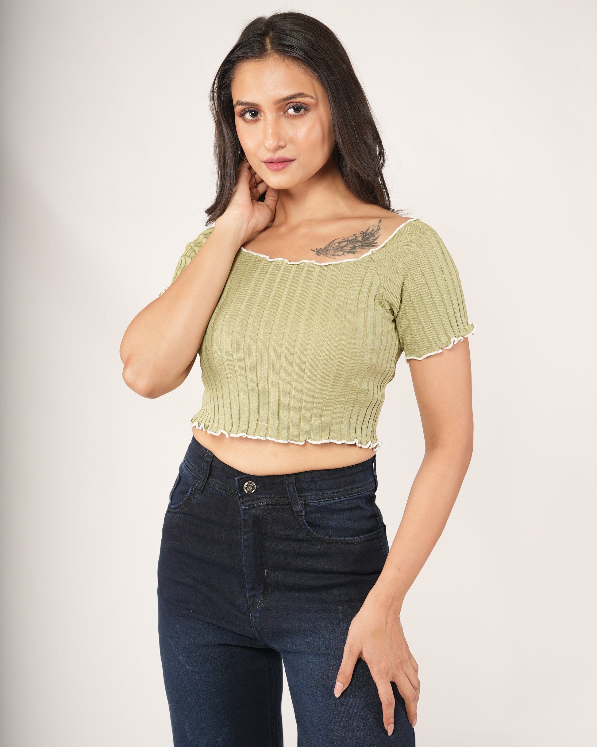 A woman modeling a light olive green ribbed short-sleeve crop top with a ruffled neckline, paired with dark blue jeans.