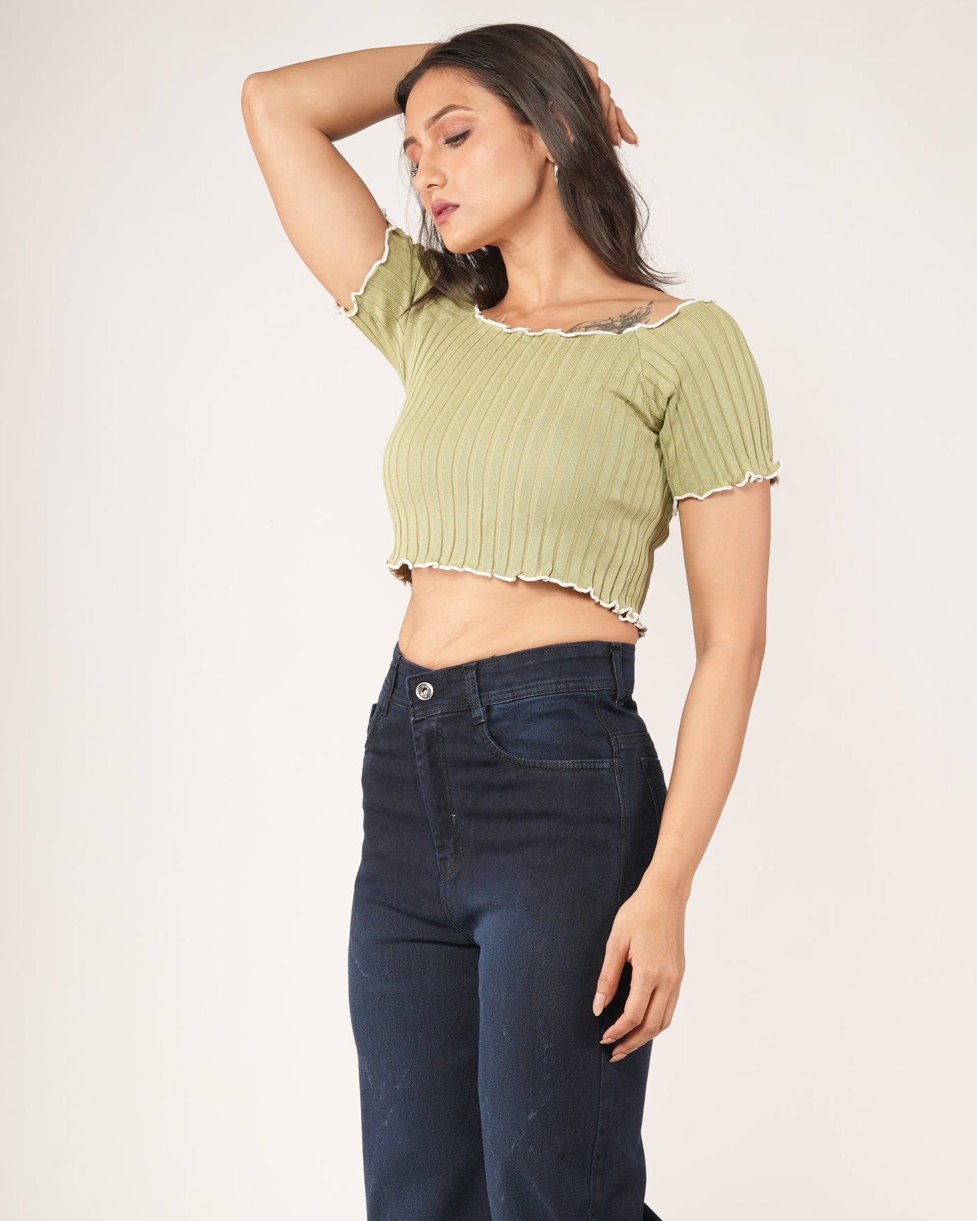 A woman modeling a light olive green ribbed short-sleeve crop top with a ruffled neckline, paired with dark blue jeans.