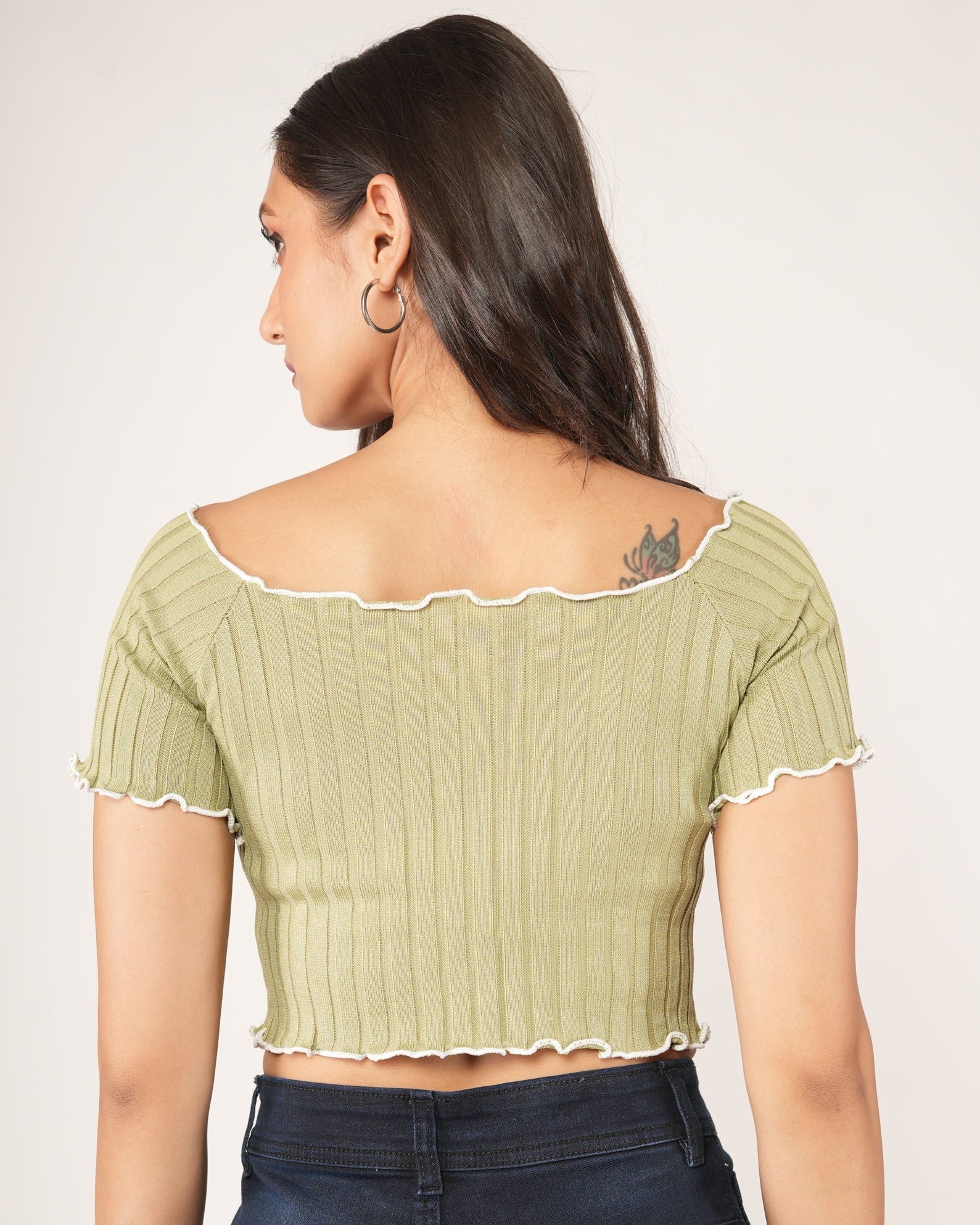 A woman modeling a light olive green ribbed short-sleeve crop top with a ruffled neckline, paired with dark blue jeans.