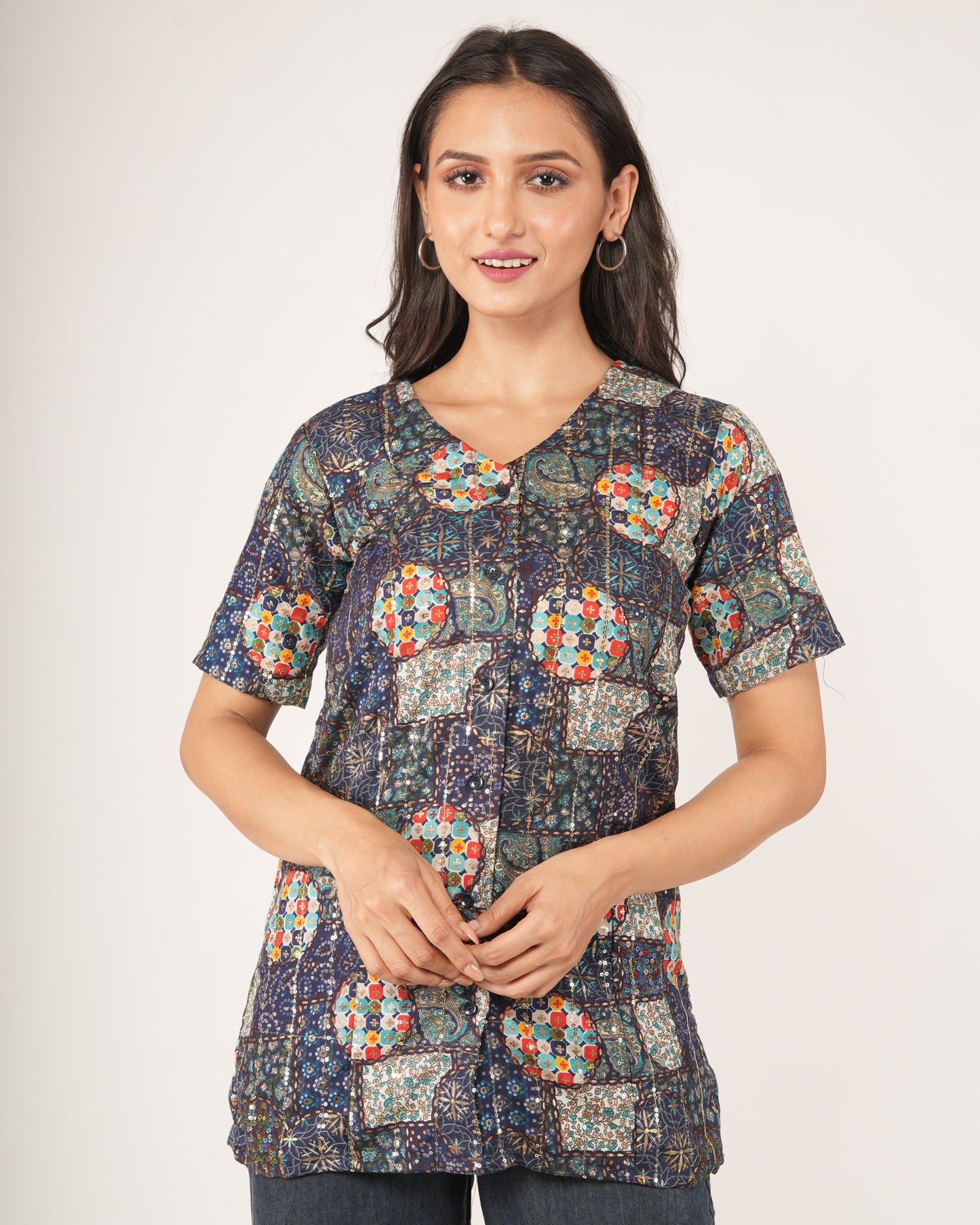 A woman modeling a short-sleeve button-up scrub top with a multicolor patchwork print on a dark navy base, paired with dark gray pants.