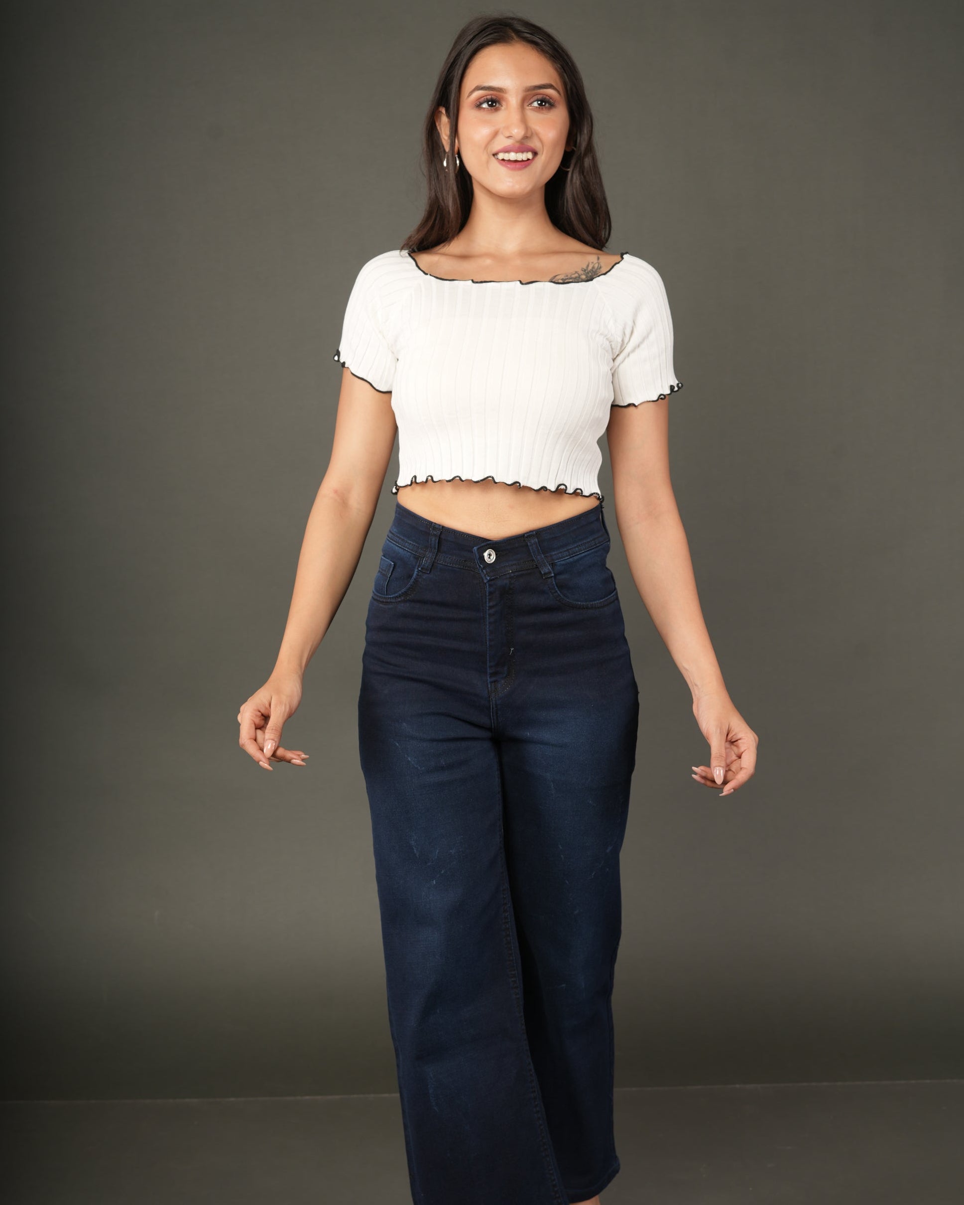 A woman modeling a white short-sleeve ribbed crop top with a scalloped black trim, paired with dark blue jeans.