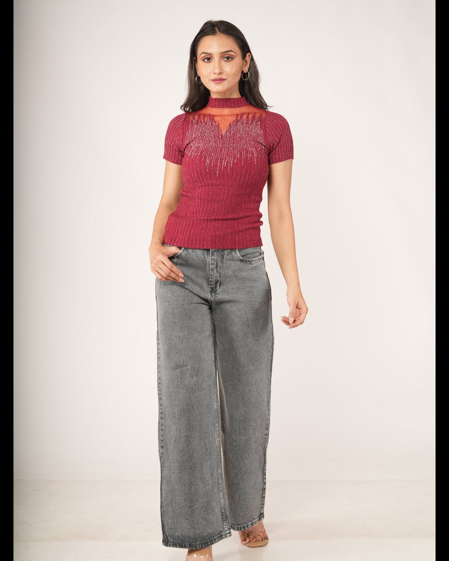A woman modeling a short-sleeve ribbed knit top in deep burgundy with a textured fringe-like pattern across the chest, paired with wide-leg gray jeans.and beige sandals.